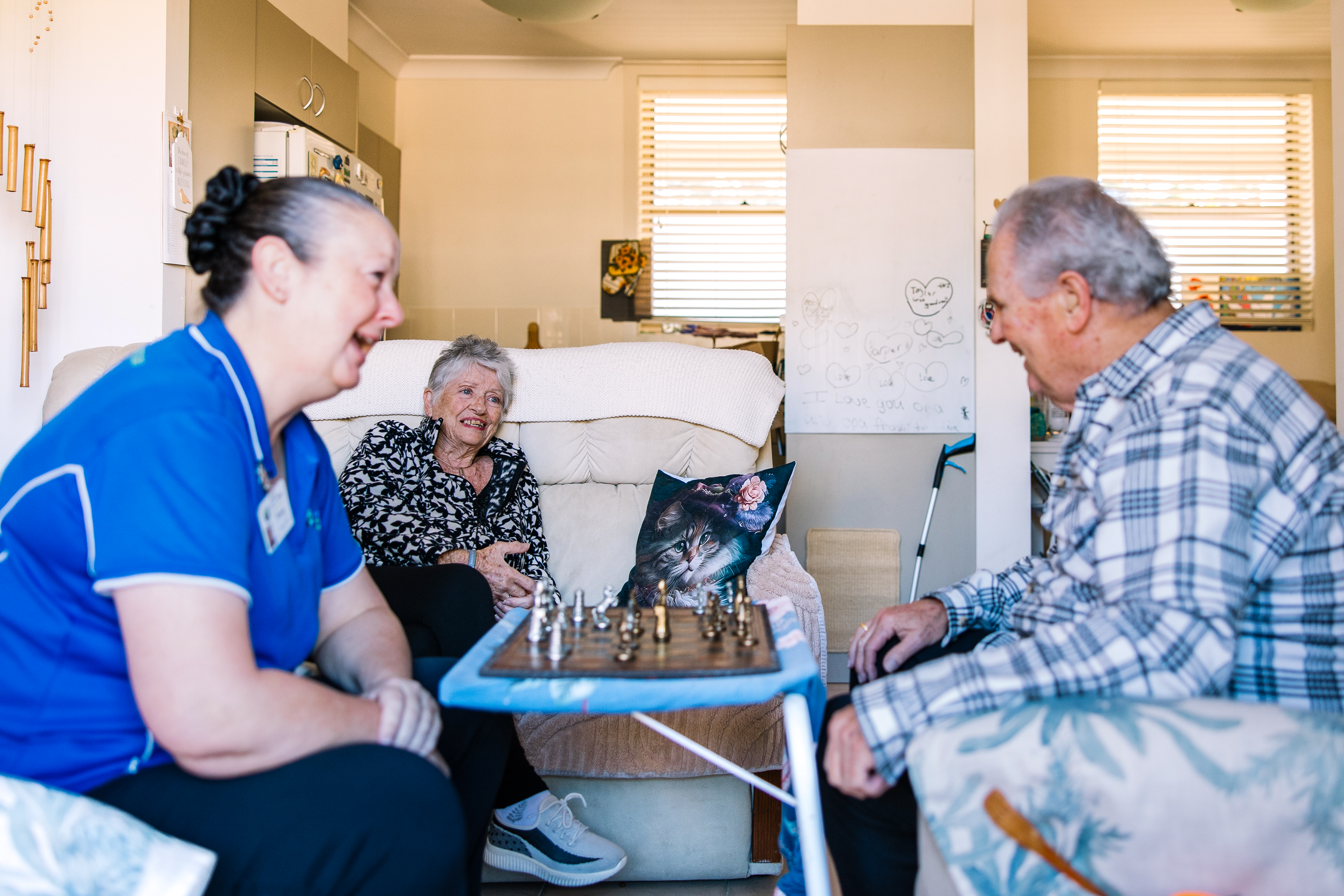 Support worker and client playing chess while a woman is seated on a couch smiling