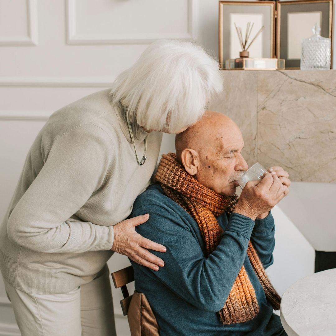 elerly woman supporting an elderly man who is drinking from a cup